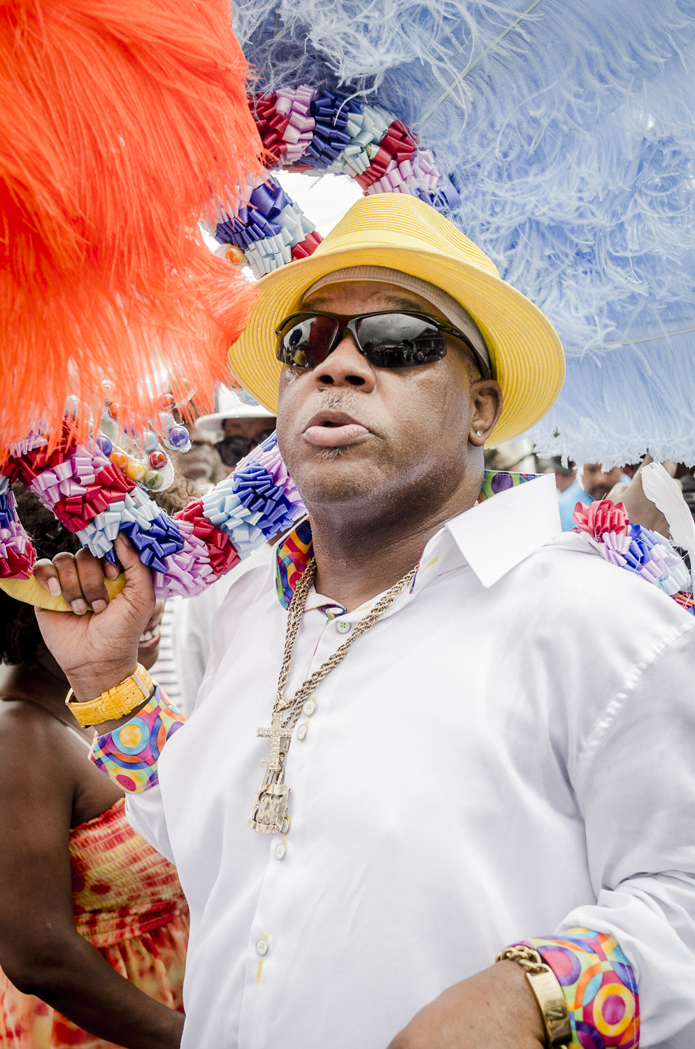 A man in a white shirt, yellow hat, and sunglasses carries a colorful feathered umbrella during a parade.