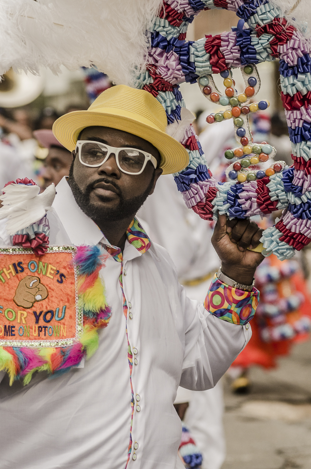 A man in a yellow hat and white outfit holds a decorated feathered umbrella and a colorful sign during a parade.