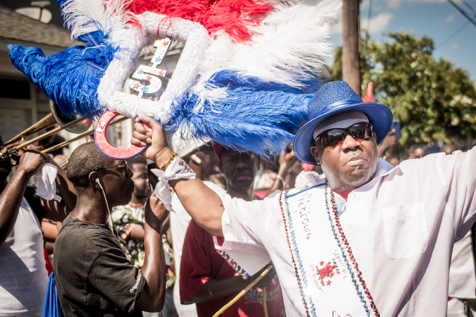 A proud second-line dancer in a white shirt, blue hat, and sunglasses energetically raises a red, white, and blue feathered accessory while leading a street parade, with a brass band playing behind him.