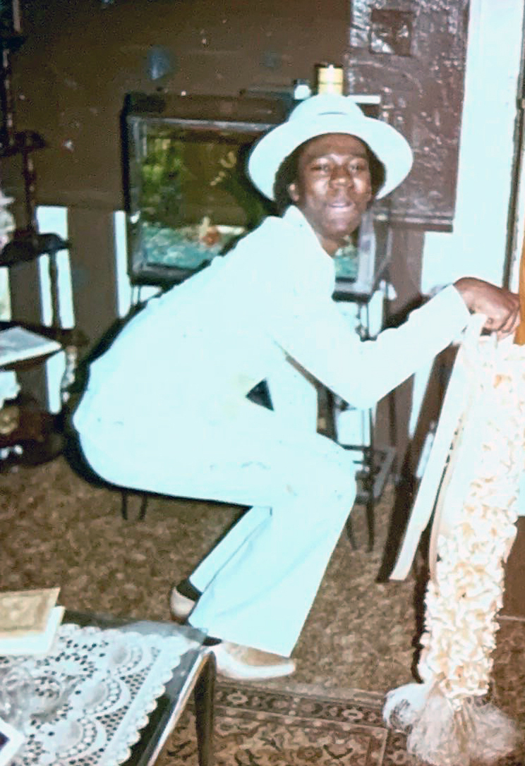 A vintage photograph of a man in an all-white suit and matching hat, striking a playful pose in a warmly decorated living room.