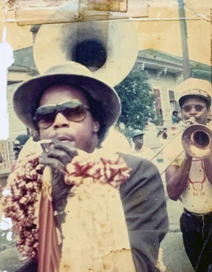 A stylishly dressed man in a dark outfit, hat, and sunglasses participates in a second-line parade, gripping an ornate umbrella as a sousaphone player follows behind him.