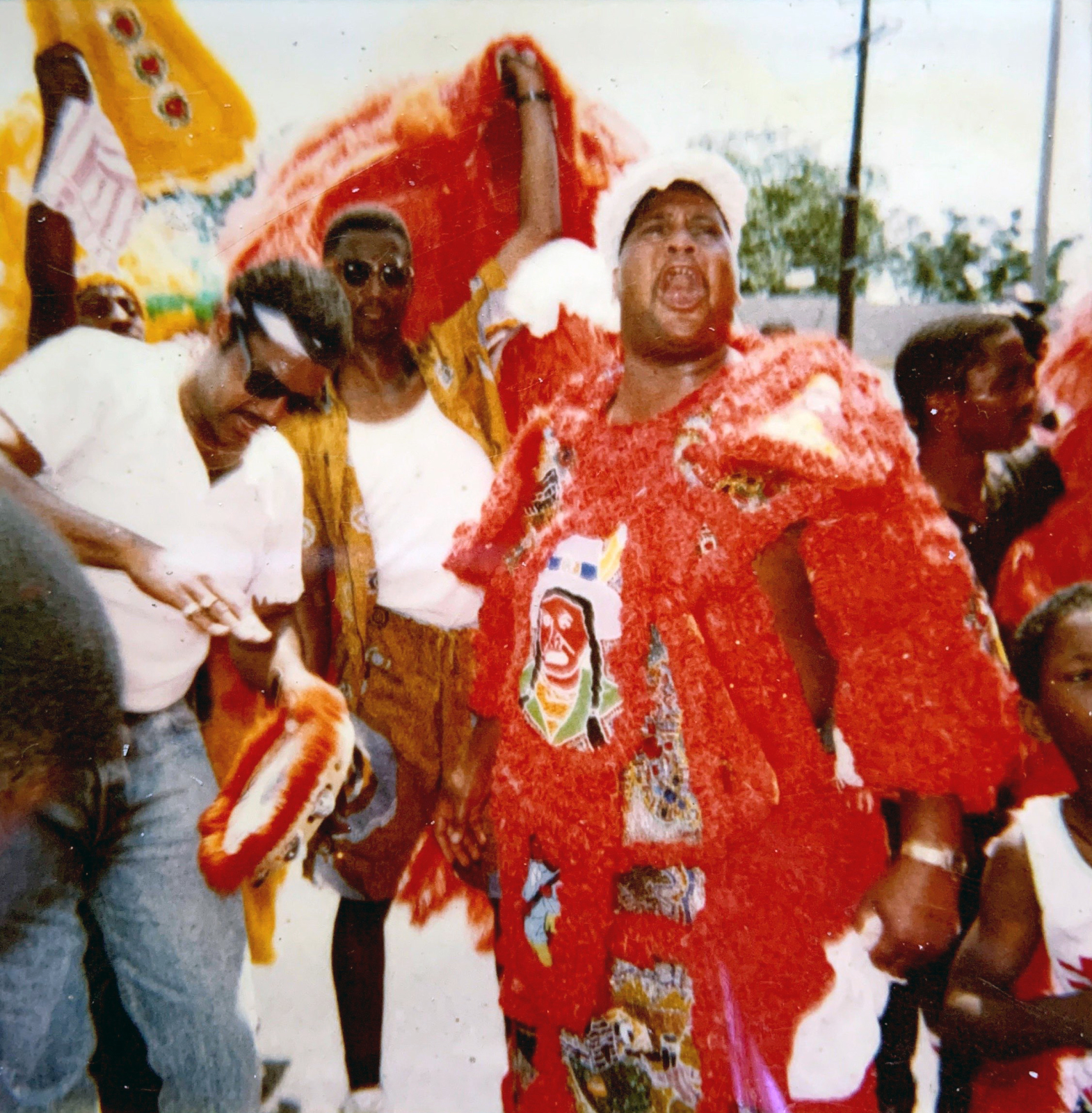 A scene from a second line parade, featuring a man in an elaborate red Mardi Gras Indian suit adorned with intricate beadwork and embroidery, passionately singing or chanting. Surrounding him are others dressed in colorful attire, some holding feathered adornments.