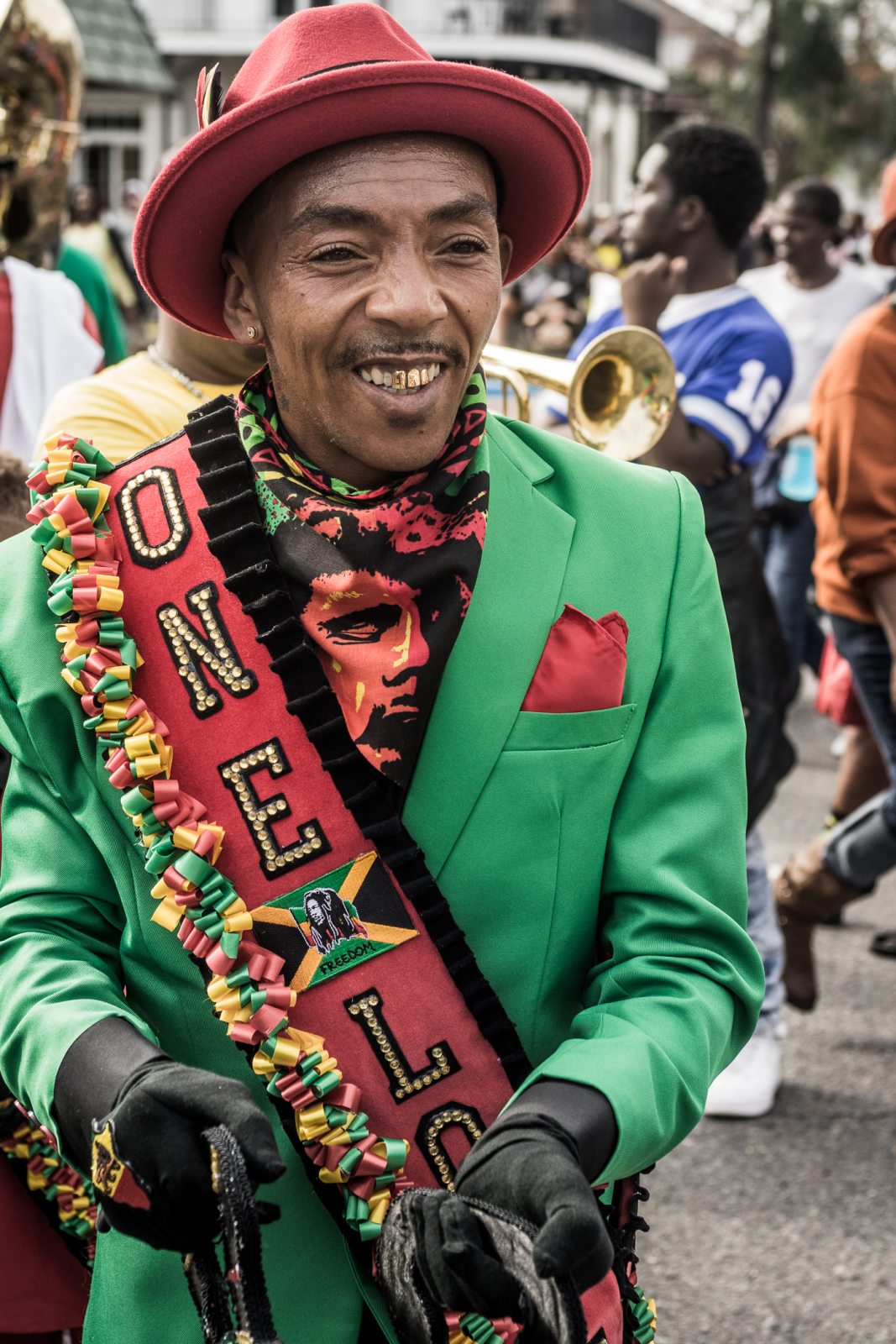A man wearing a vibrant green suit, red hat, and black gloves smiles as he marches in a parade, adorned with a sash reading "ONE LOVE" in red, green, and yellow, while a brass band plays behind him.