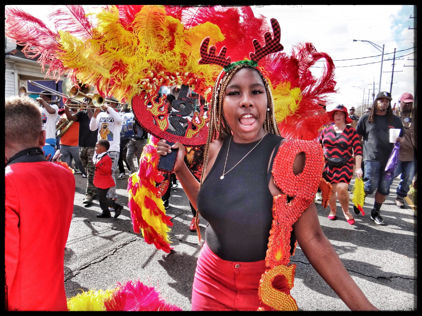 A young woman in a black and red outfit, wearing a festive antler headband, marches with enthusiasm, holding an ornate red and black fan decorated with feathers as a brass band and parade-goers celebrate behind her.