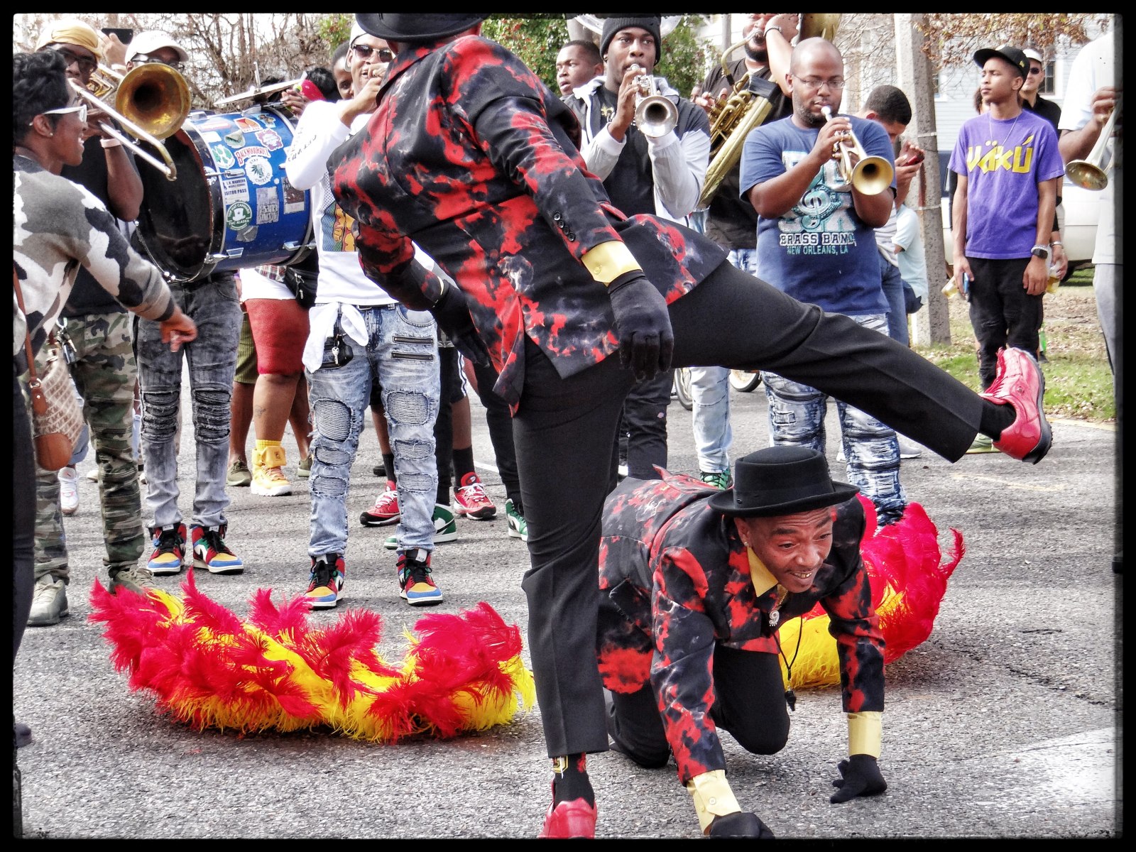 A lively parade scene captures two dancers mid-performance, dressed in coordinated red and black attire, with one bending low as the other leaps over, while a brass band plays in the background.