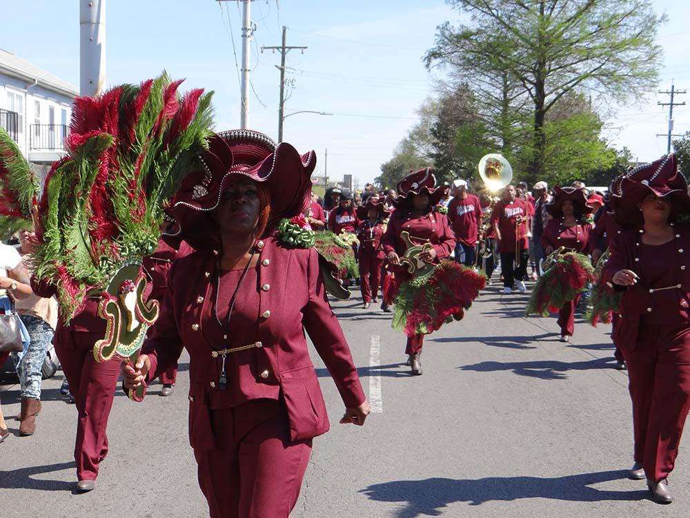 A group of people in matching maroon outfits and large hats adorned with green feathers marches down a street during a parade. They hold decorated umbrellas and dance energetically. In the background, a brass musician follows.