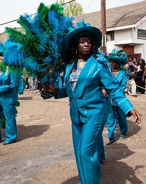 A person dressed in a vibrant blue suit and hat participates in a parade, holding a large blue and green feathered fan. They are marching outdoors, with houses and a crowd visible in the background.