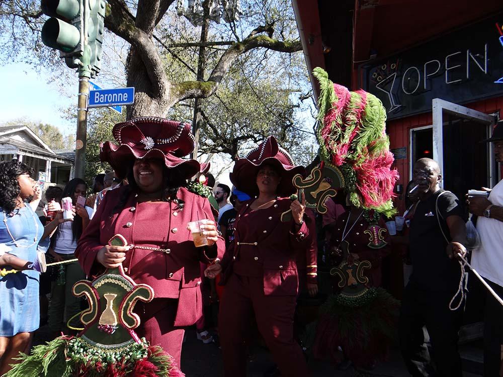 A lively street scene in a festive parade with participants wearing matching burgundy outfits and elaborately decorated hats, holding vibrant green and red parasols. Onlookers watch under trees, and a street sign reads Baronne.