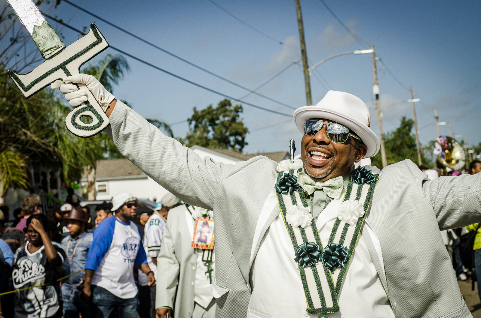 A joyful man in a light green suit and white hat spreads his arms, holding a decorated "9"-shaped staff during a parade.