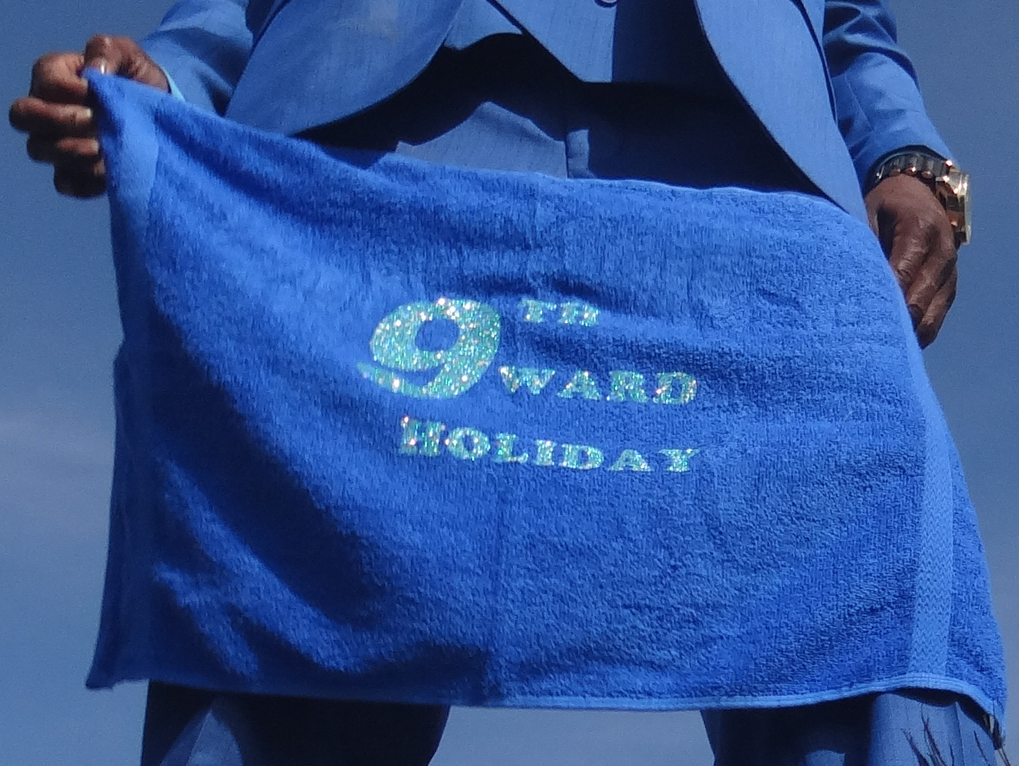 A person in a blue suit holds a blue towel with glittery text that reads "9th Ward Holiday."
