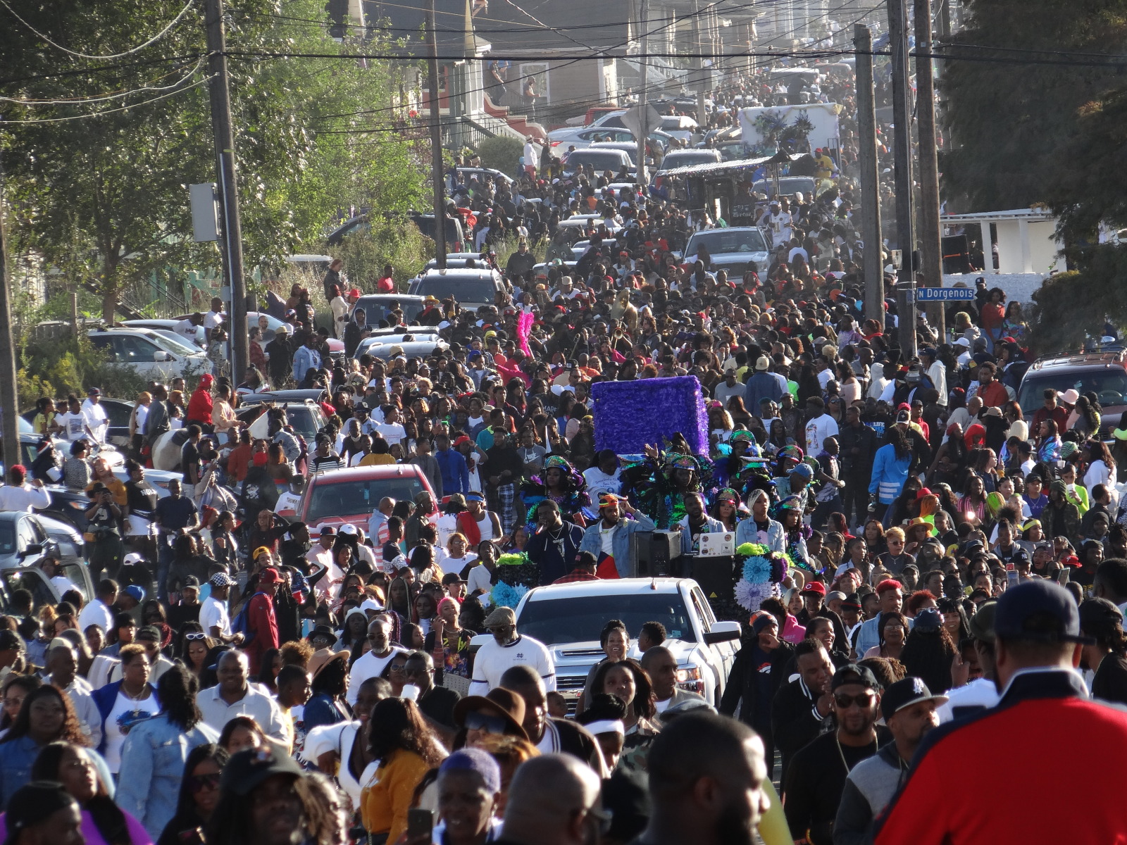 A lively street parade with hundreds of people in vibrant clothing, some wearing feathered headdresses, while cars and trucks navigate through the packed roadway.