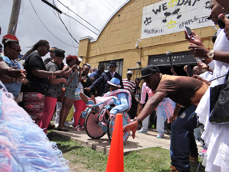 A lively street scene with a crowd of people gathered around a person in colorful attire performing stunts on a bicycle. Some are taking photos, while others cheer. A banner reading BEAUTIFUL PEOPLE LLC is visible in the background.