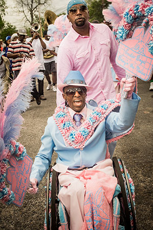 A joyful person in a wheelchair, dressed in a colorful blue and pink costume with elaborate decorations and feathers, takes part in a parade. A group of people in festive clothing and musicians can be seen in the background.