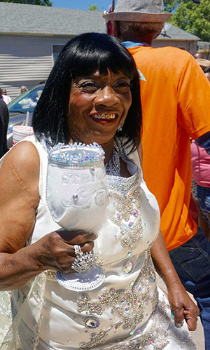 Elderly woman smiling, wearing a white embellished dress and holding a bejeweled goblet. She is outdoors, standing next to someone in an orange shirt, with houses and trees in the background.