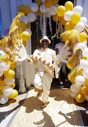 A person in a festive ivory and gold outfit dances energetically, holding gold and white feathered fans. They are surrounded by a decorative archway of white and yellow balloons, on a path lined with a gold carpet.