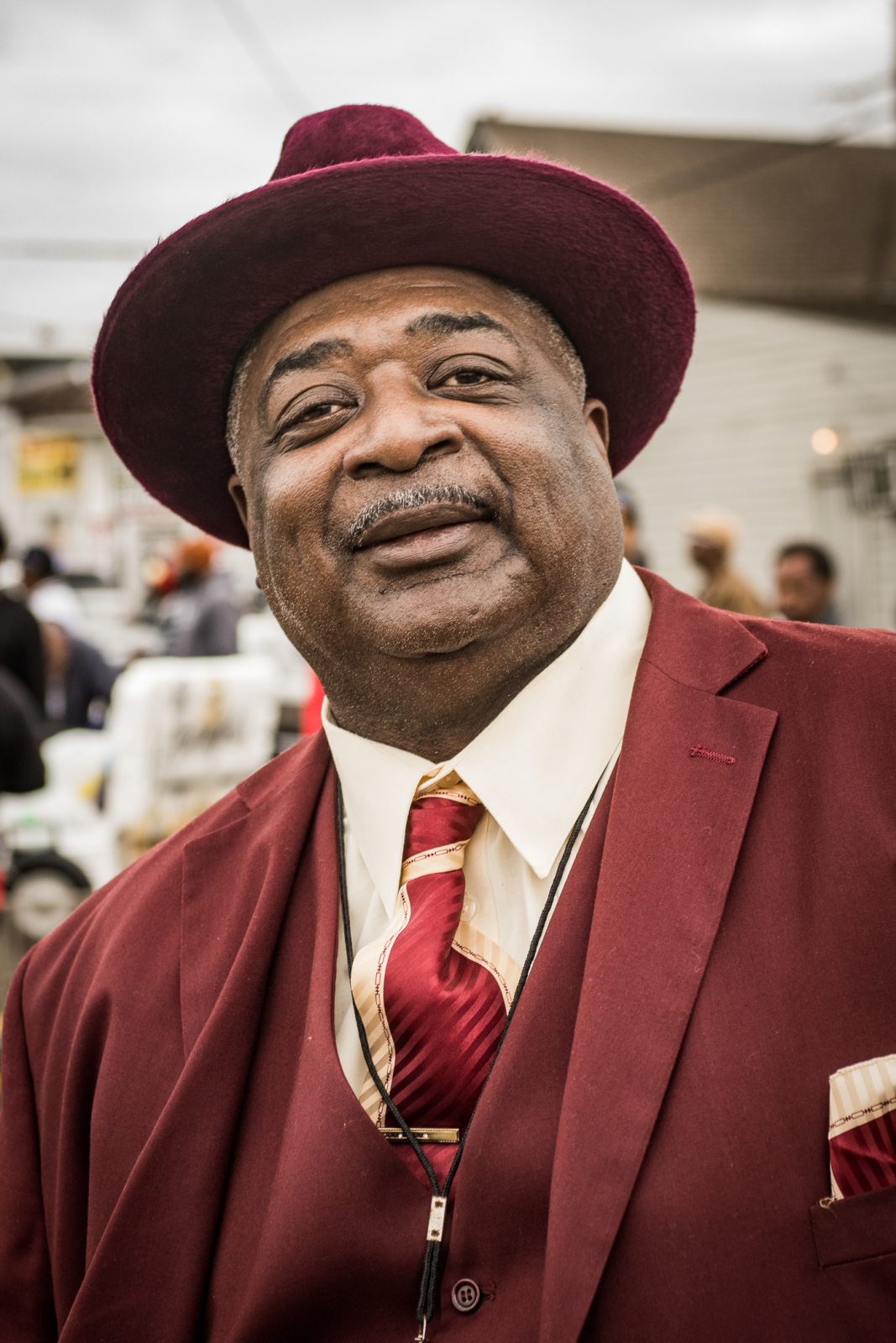 Dressed in a sharp burgundy suit and hat, a man exudes confidence with a relaxed smile and a stylishly knotted tie.