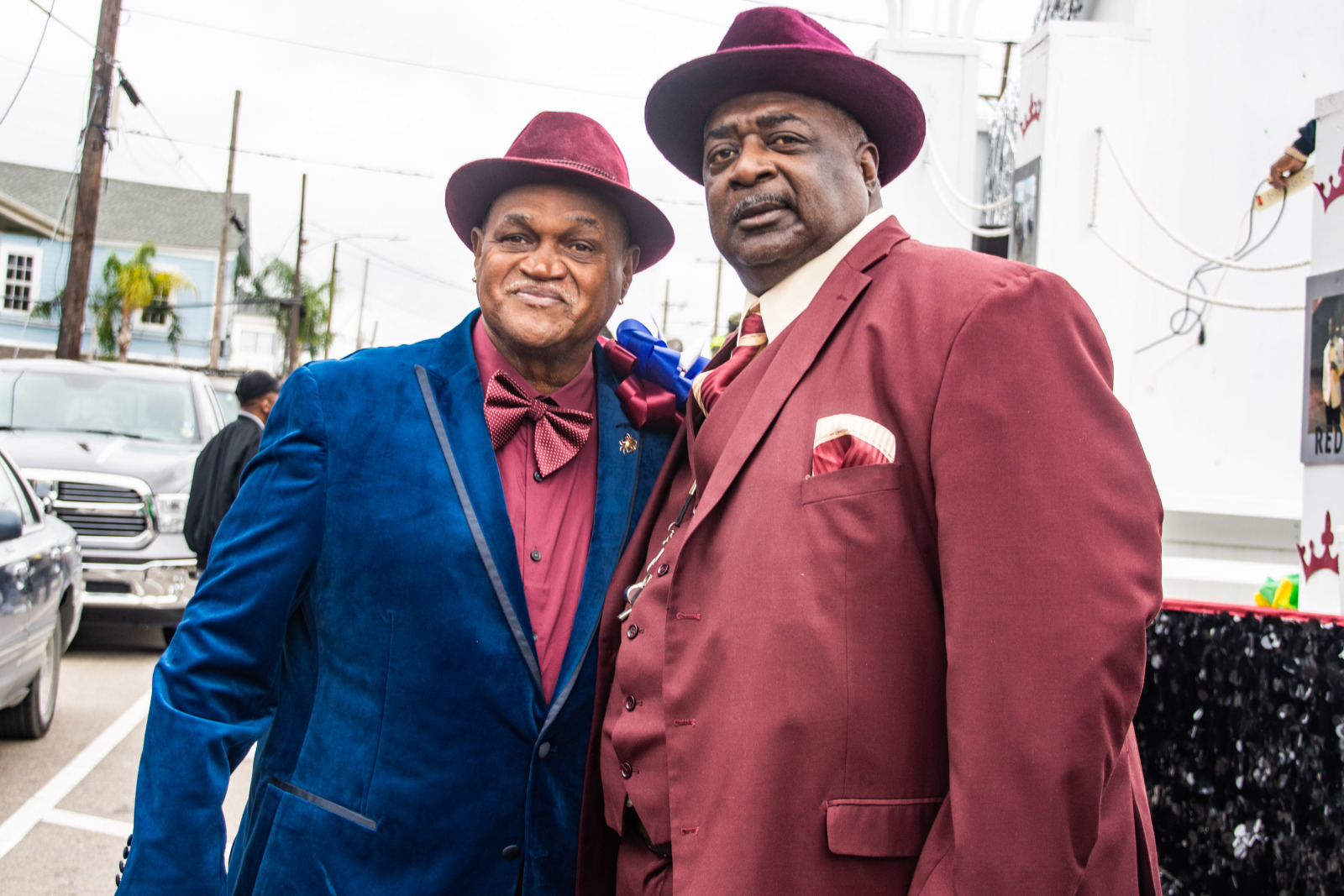Two men in sharp suits and matching hats pose together, one in deep burgundy and the other in rich blue, exuding style and confidence.