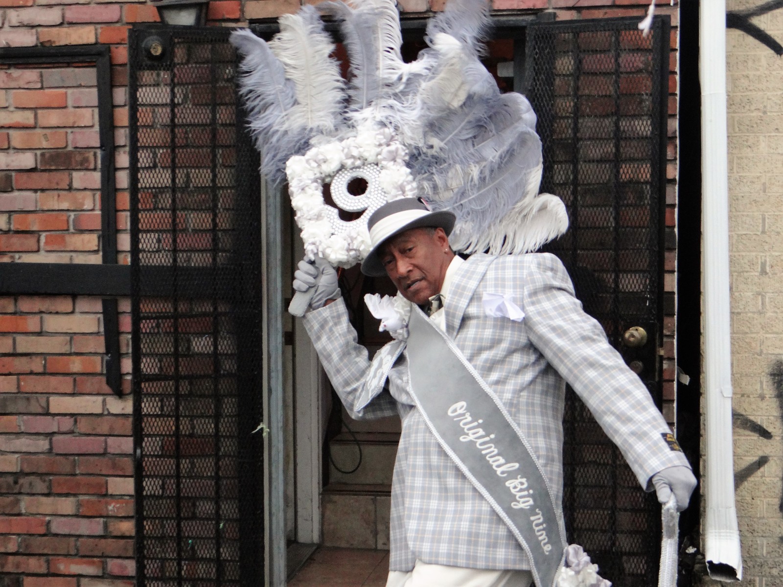 A sharply dressed man in a light gray plaid suit and matching hat strikes a pose, holding an elaborate feathered fan with "Original Big Nine" written on his sash.