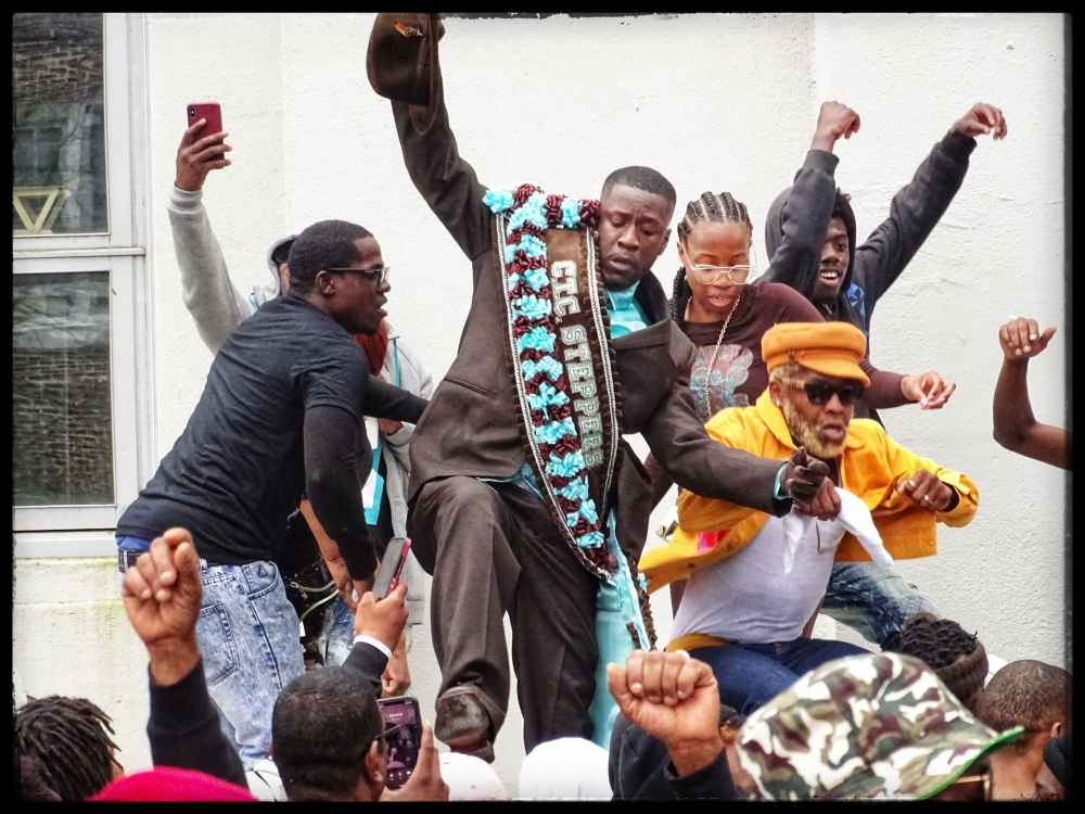 A group of people celebrating energetically, with one person in focus wearing a suit and a sash that reads CLC Spartans. Others around them are cheering and taking photos, with raised fists and vibrant expressions.