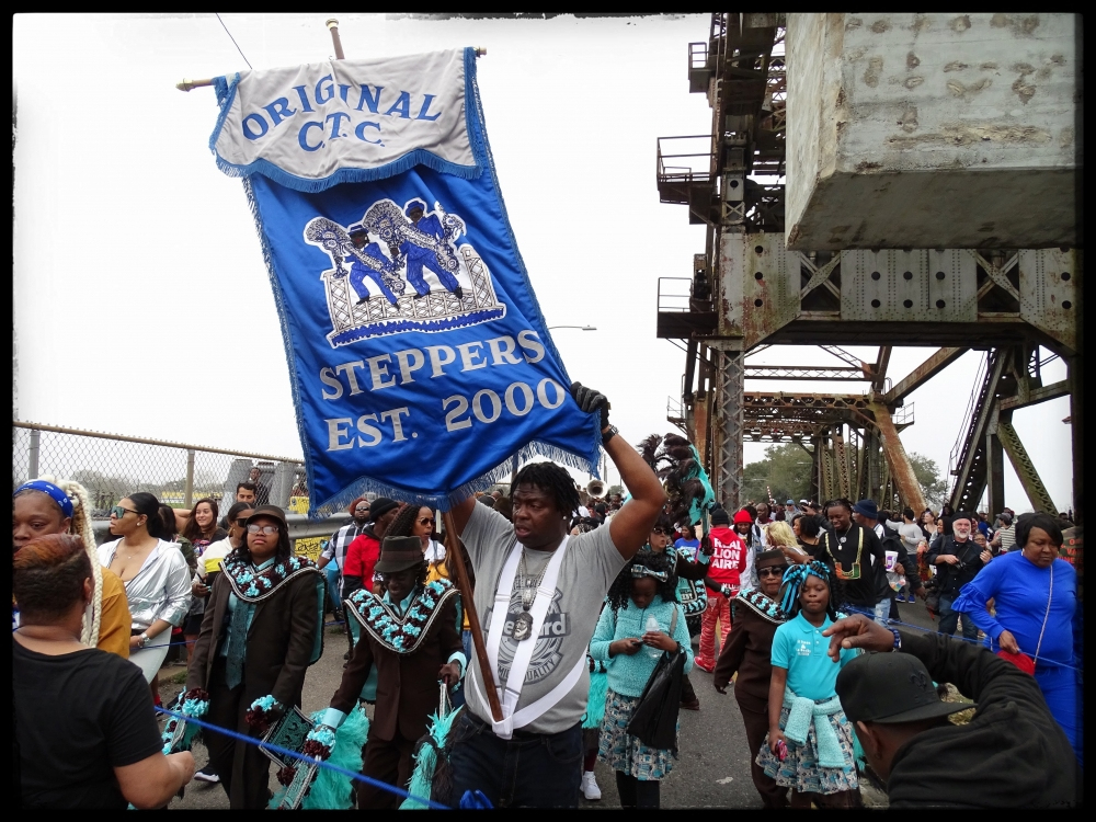 A group of people parades on a bridge under a blue banner that reads Original C.T.C. Steppers Est. 2000. They are dressed in colorful outfits, some with feathered accessories, amidst a lively crowd. Theres industrial metalwork in the background.