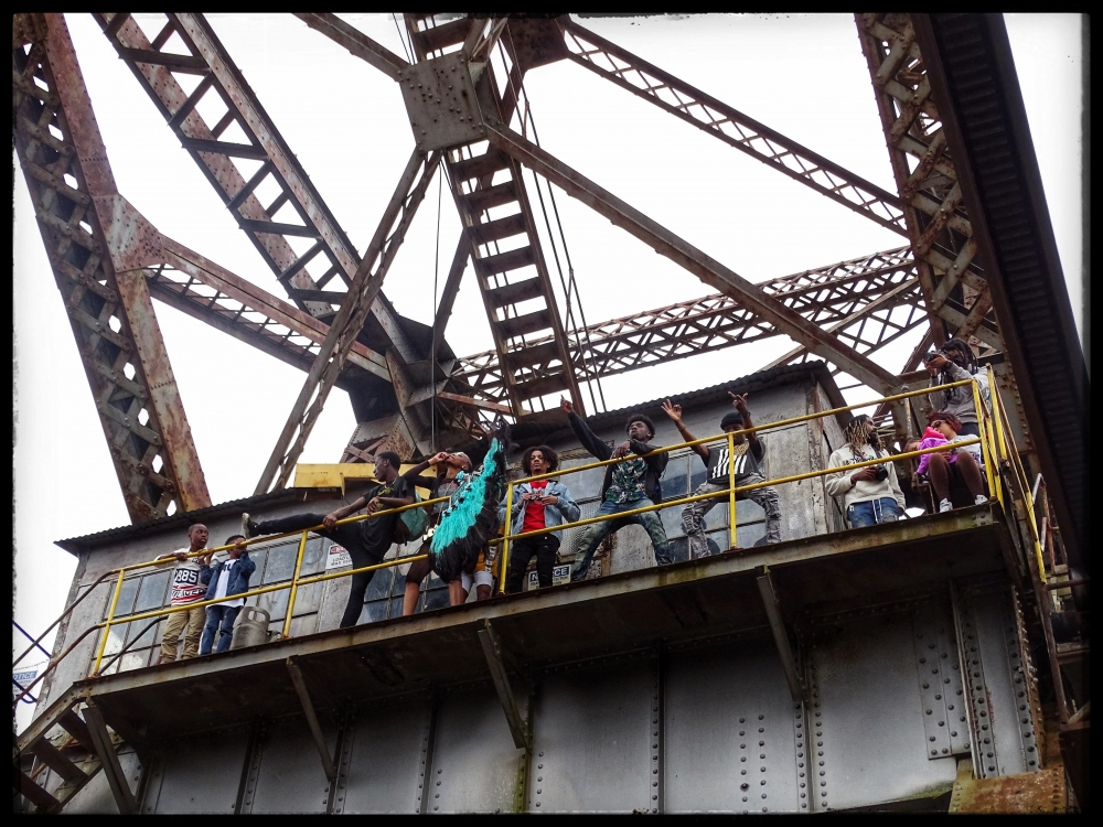 A group of people stands on a metal platform under a rusty steel structure. Some are waving, and one holds a blue flag. The scene appears industrial, with beams and girders overhead.