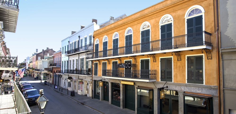 Street view of a charming historic city with colorful buildings. The architecture features arched windows and wrought-iron balconies. The street is quiet, with a few parked cars, under a clear blue sky.