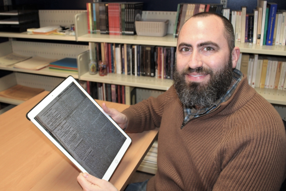 A bearded man in a brown sweater sits at a desk in a library. He smiles at the camera while holding a tablet displaying a document. Bookshelves filled with books are in the background.
