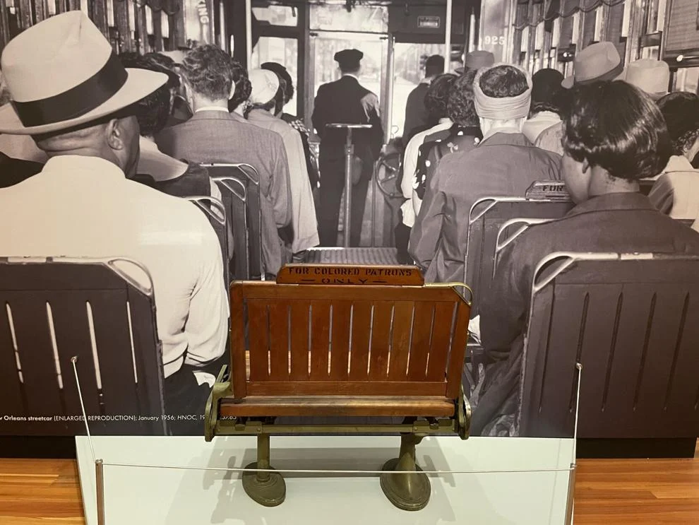 An exhibition installation shows a streetcar bench from a New Orleans streetcar, labeled "FOR COLORED PATRONS," and placed in front of a vintage black and white photo of a streetcar from the same era.