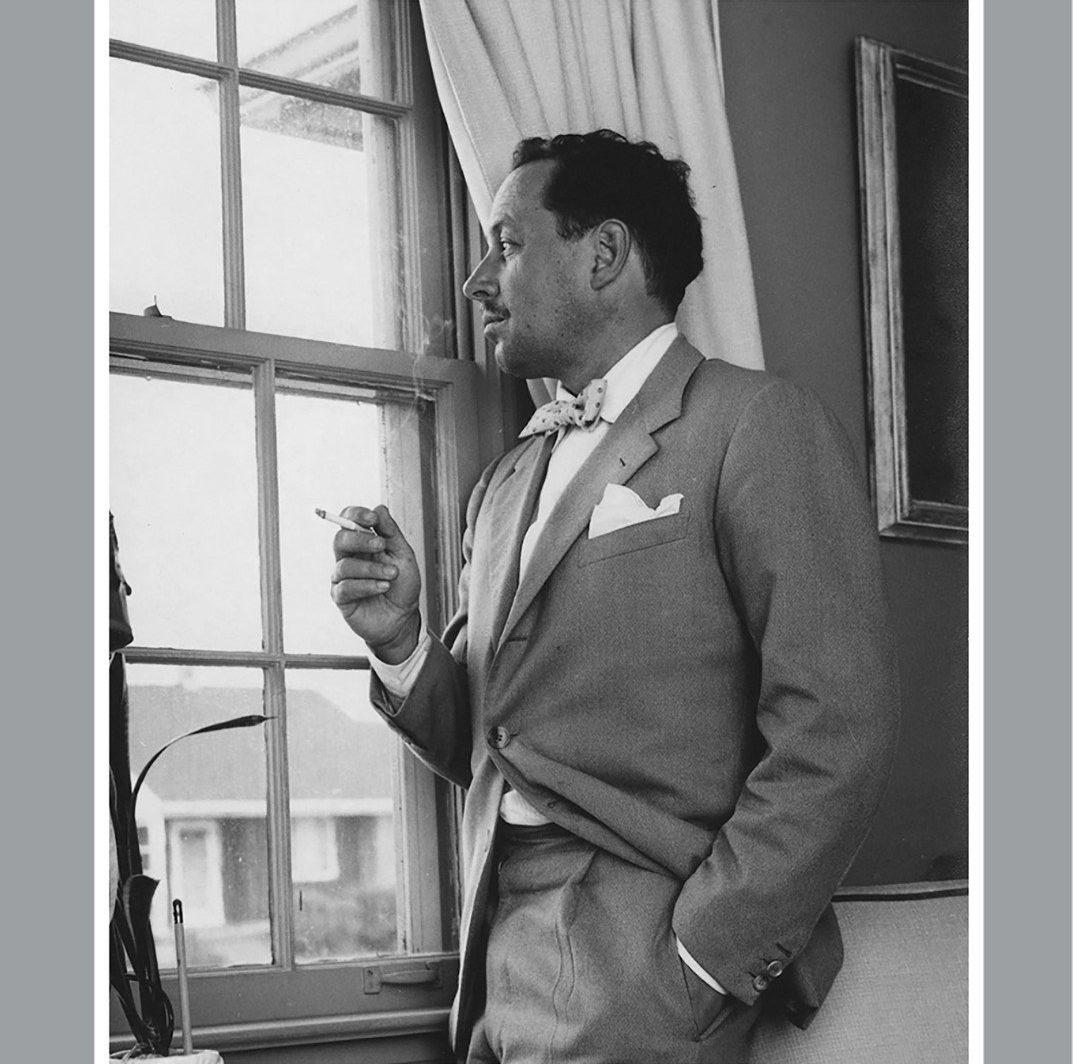 A person in a suit and bow tie stands indoors, holding a cigarette and looking out a window with curtains. The room has a picture frame and a potted plant visible in the background.