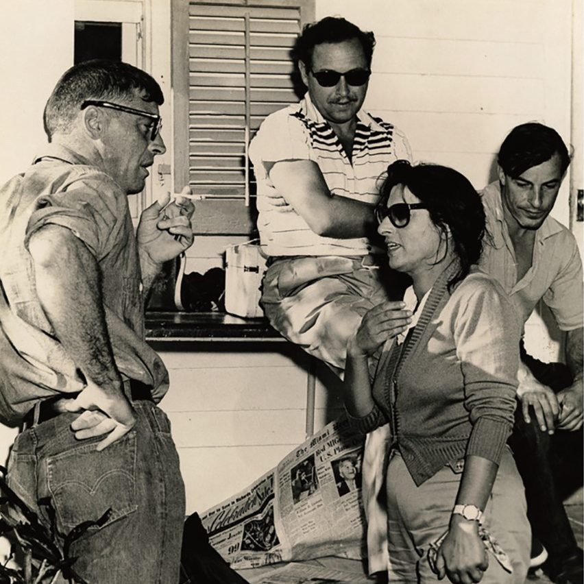 A vintage black-and-white photo of four adults having a casual conversation outdoors. Two men are leaning against a wall, one sitting on a windowsill, and a woman stands talking. Newspapers are spread out nearby.