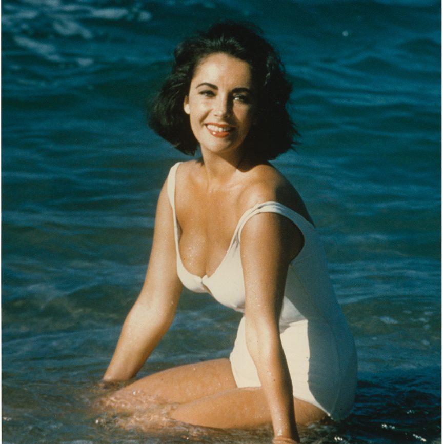 A woman in a white swimsuit kneels in shallow water at the beach, smiling at the camera. The ocean waves gently surround her.