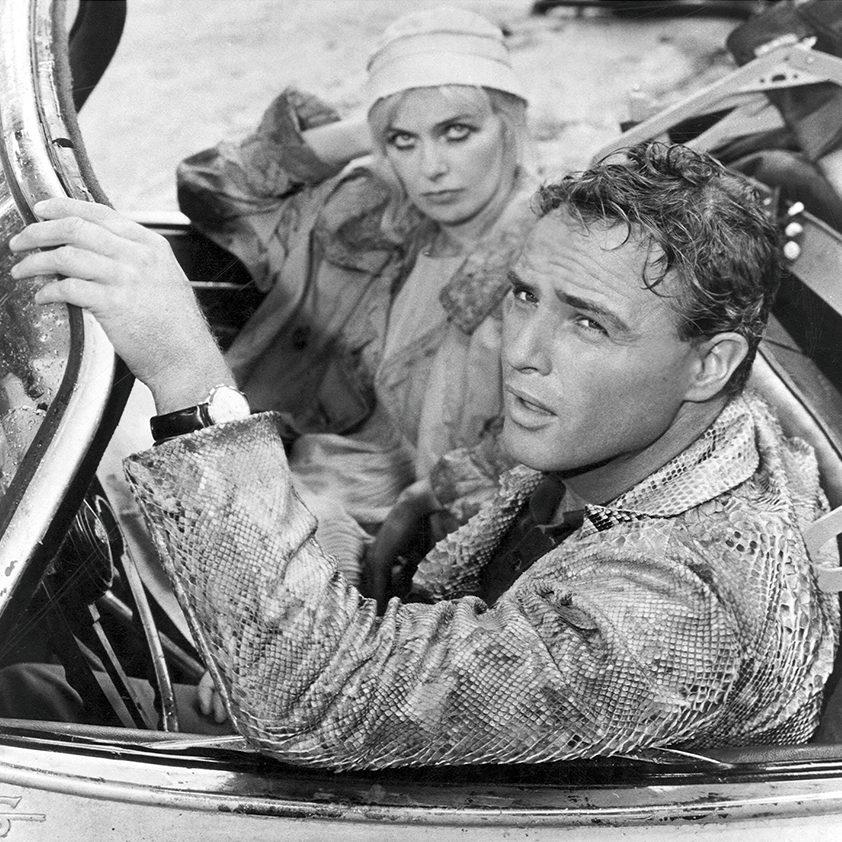 A black and white photo of a man and a woman sitting in a convertible car. The man, in the drivers seat, gazes outward, wearing a textured jacket. The woman, seated next to him, has a headscarf and coat, looking subtly towards the camera.