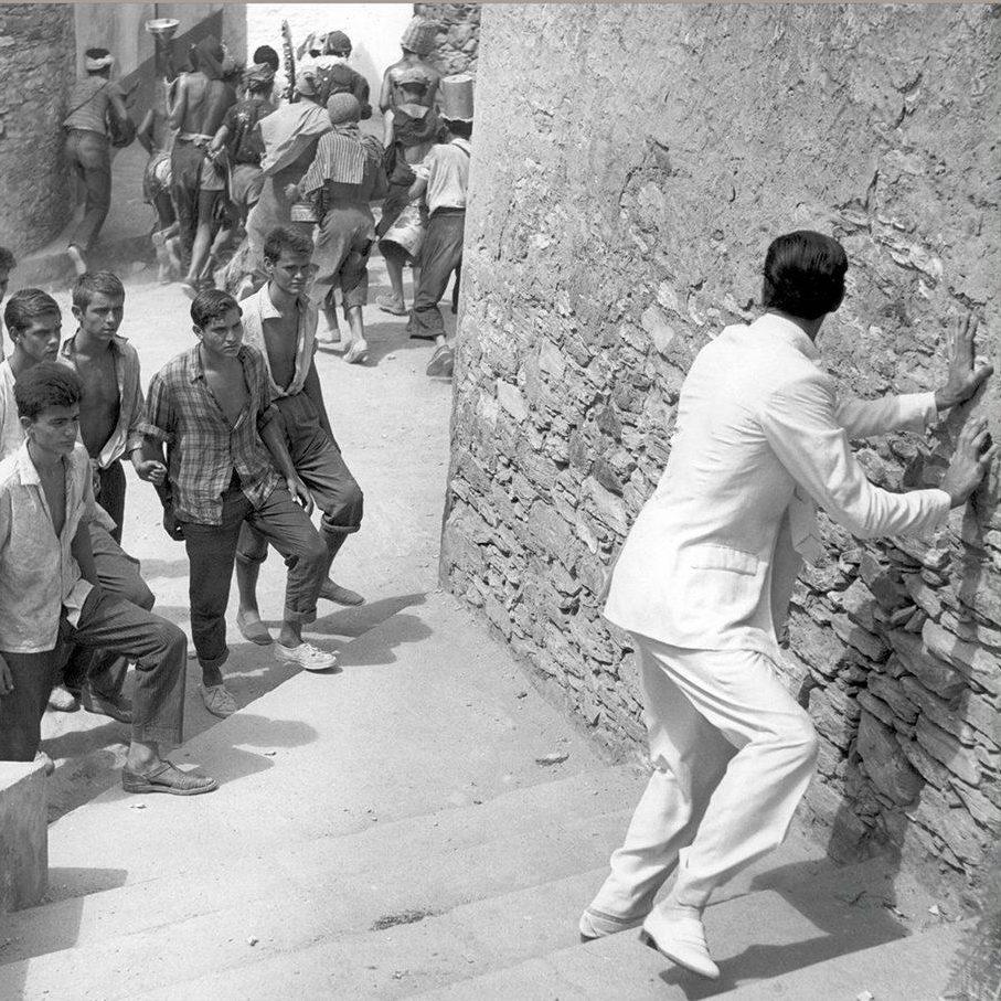 A man in a light suit climbs a narrow stone staircase with a group of casually dressed young men following behind. They ascend in an outdoor area surrounded by stone walls.