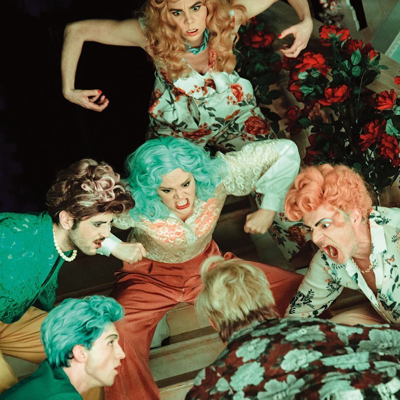 A dramatic scene with five people in colorful costumes and wigs, gathered on a staircase. They appear to be in an intense, theatrical argument. The setting is vibrant with floral decorations, enhancing the dynamic atmosphere.