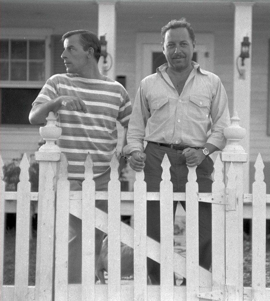 Two men stand behind a white picket fence in front of a house. The man on the left wears a striped shirt and looks to the side, while the man on the right wears a button-up shirt and smiles at the camera. The house has a porch with visible windows and doors.