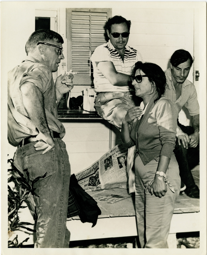 A group of four people socialize on a porch, with three men and one woman. One man sits on a railing, the others stand or lean casually. A newspaper lies on a nearby bench. The setting appears relaxed and informal.