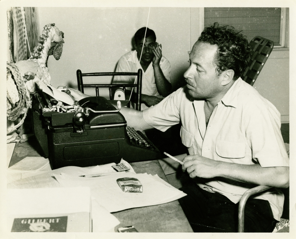 A man sits at a desk, typing on a typewriter. Papers and a pack of cigarettes are visible. Another person is seated in the background. The room appears casual, with various objects around.