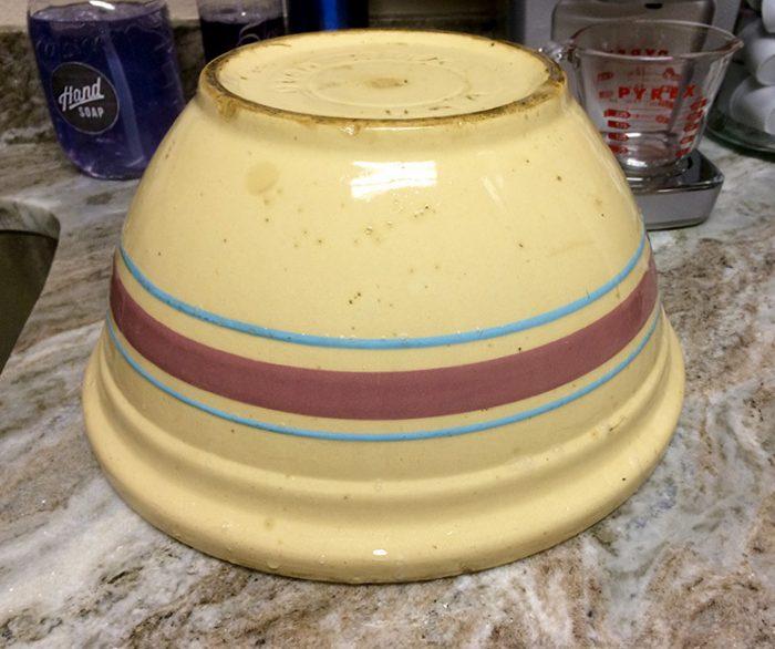 A vintage yellow ceramic bowl is turned upside down on a kitchen counter. It has a maroon and two blue stripes around its side. In the background, theres a measuring cup and some kitchen items.