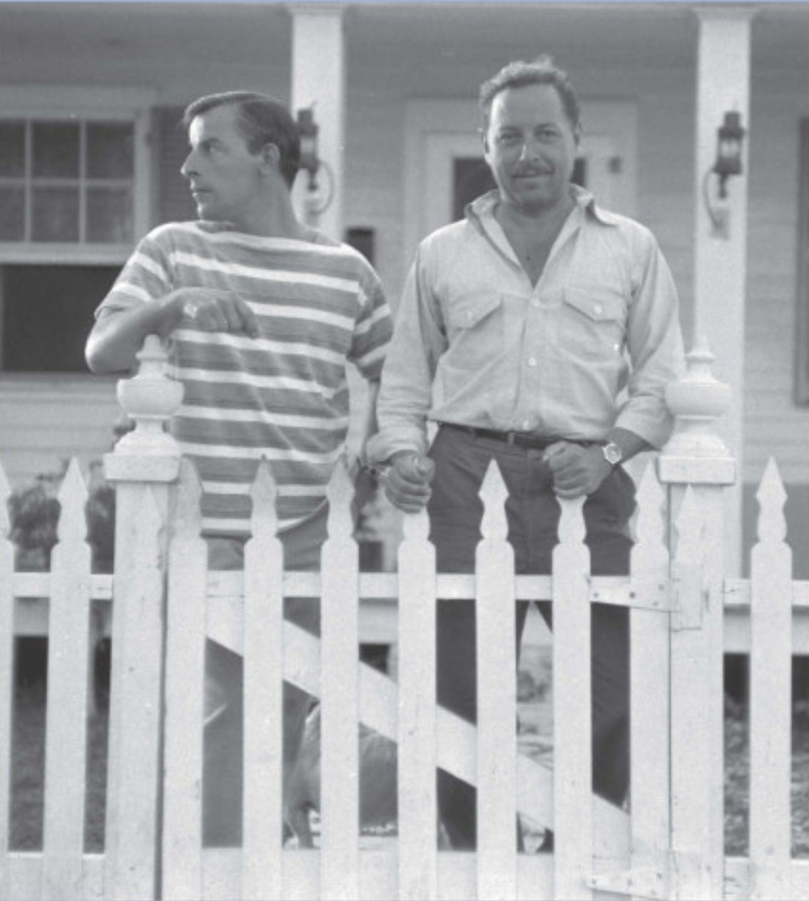 Two men stand behind a white picket fence in front of a house. One wears a striped shirt, while the other is in a button-up. They appear relaxed and are looking in opposite directions. The house has a porch and windows.
