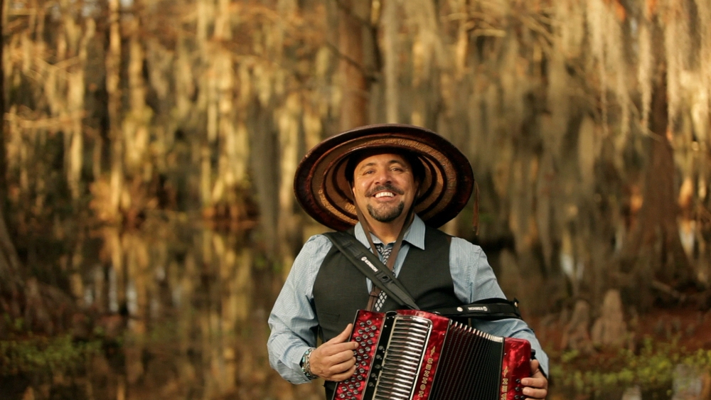 A man wearing a large brimmed hat and a vest over a blue shirt smiles while playing an accordion. He is standing in a lush, outdoor setting with trees and hanging moss in the background.