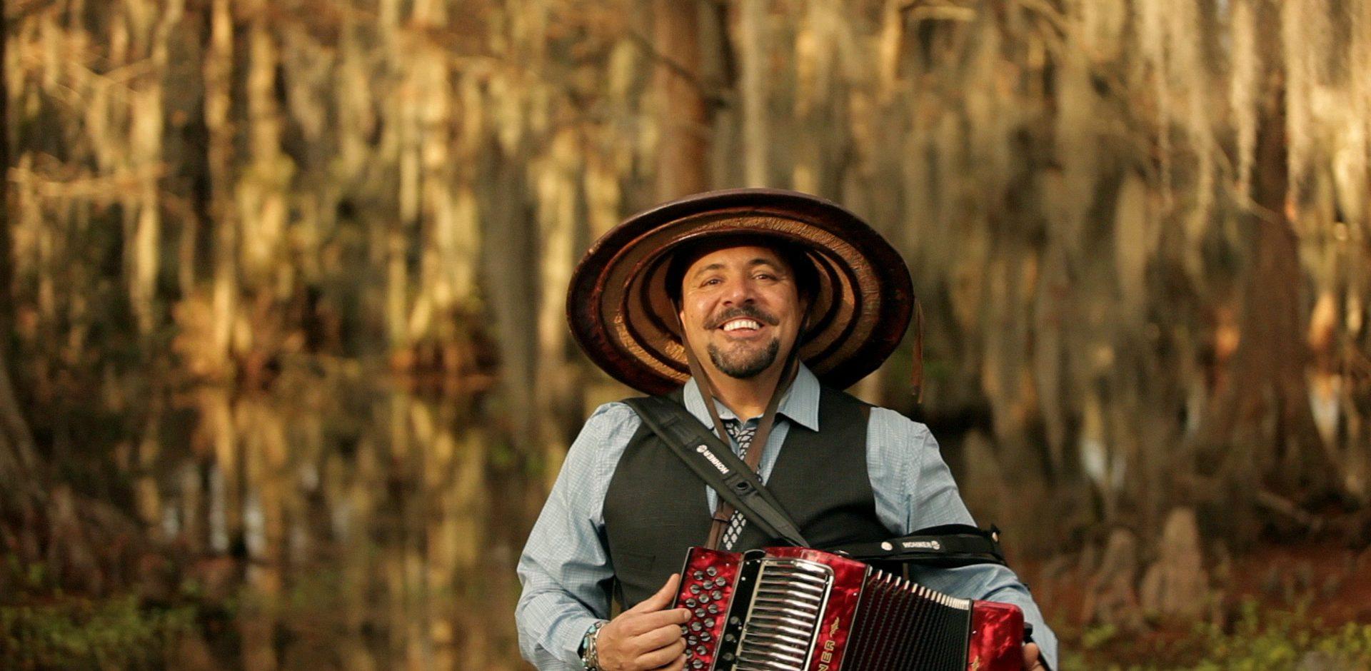 A man smiling and playing an accordion outdoors. He is wearing a large woven hat and stands in front of a backdrop of trees draped with Spanish moss.
