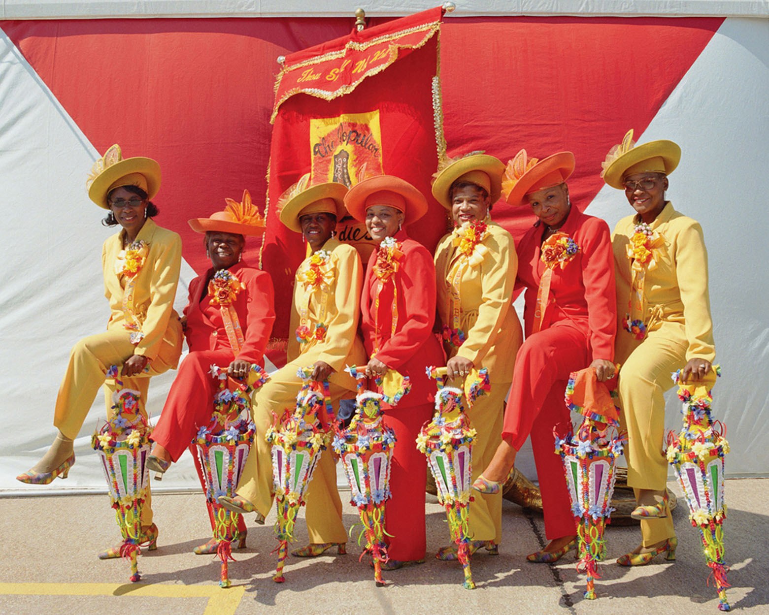 A group of eight people dressed in bright red and yellow suits and hats stand in front of a large red banner. They hold colorful umbrellas adorned with flowers and ribbons, posing together in a cheerful, celebratory manner.