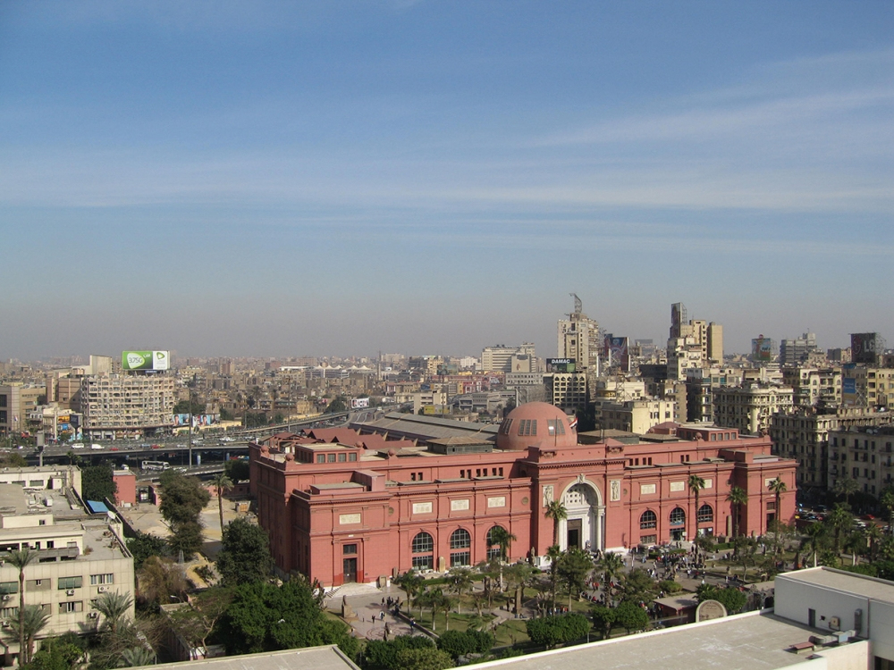Aerial view of a large, red-colored building with a dome and arched entrance, surrounded by trees and people. The cityscape in the background features various modern buildings under a clear blue sky.
