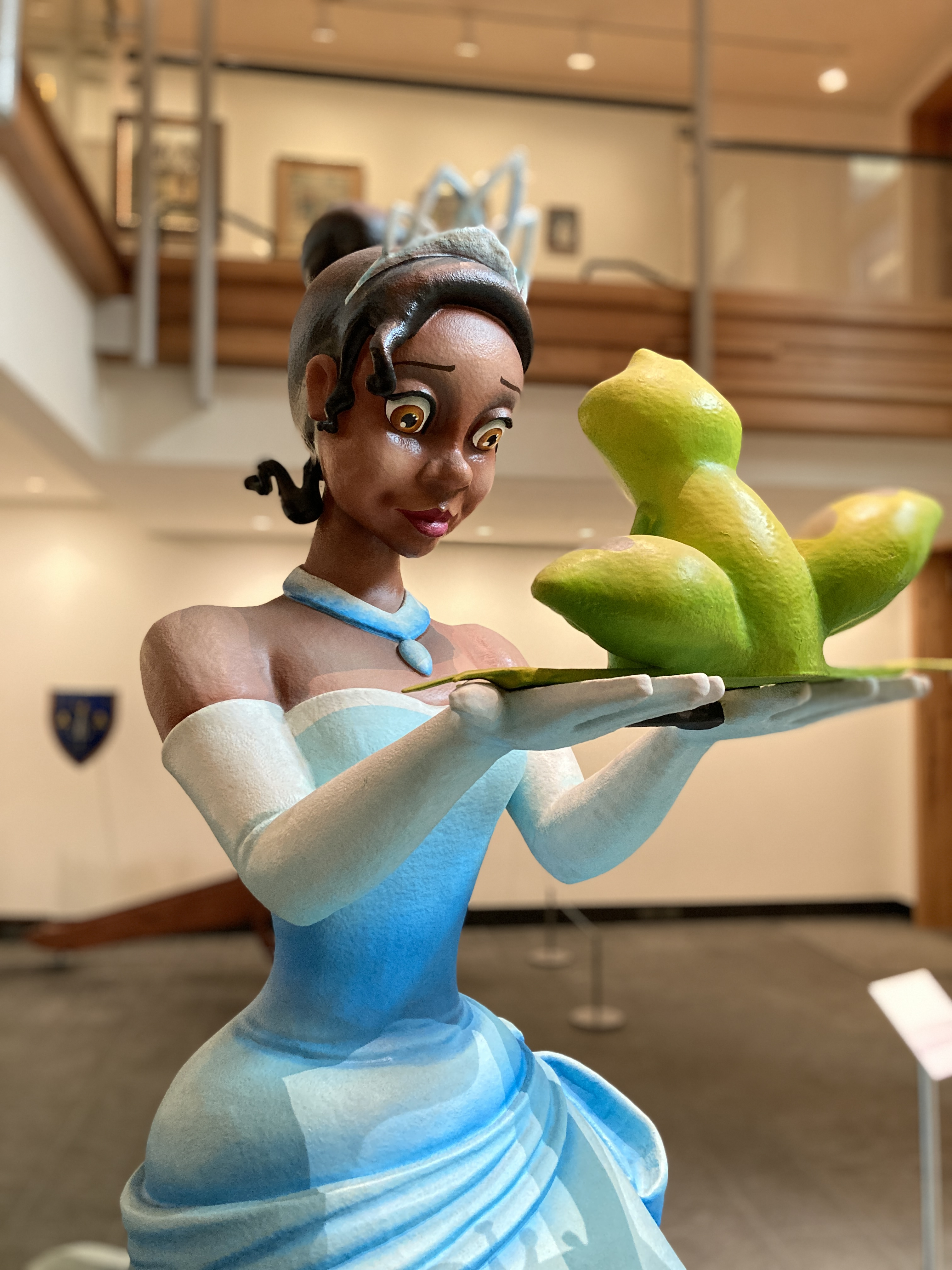 A sculpture of a woman in a blue dress and tiara holding a green frog on a plate. The setting appears to be an indoor exhibition space with wooden railings and shield-shaped decor on the walls.