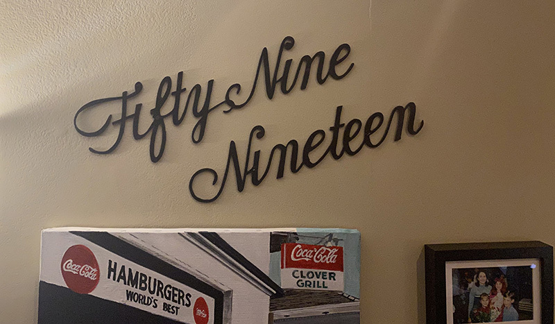 Wall decor with the words Fifty Nine Nineteen in cursive. Below, a vintage sign reads Hamburgers Worlds Best with a Coca-Cola logo and Clover Grill. A framed photo is partially visible to the right.