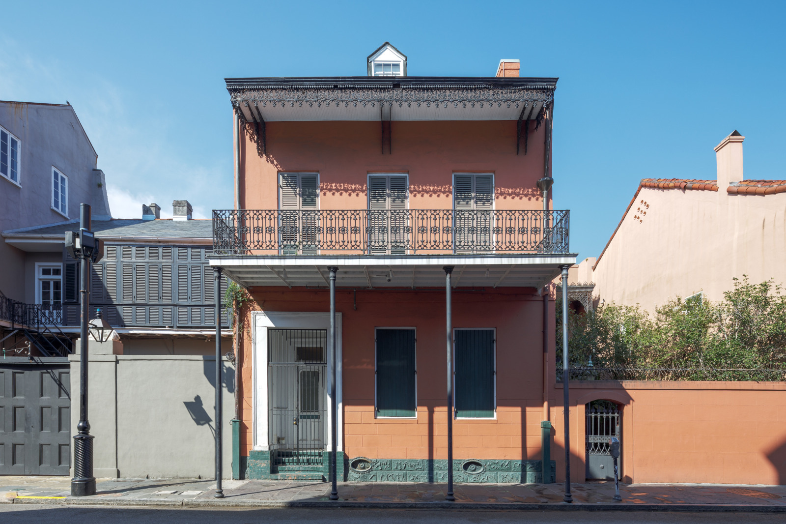 A historic two-story building with a peach facade and ornate black wrought-iron balcony in a sunny street. The building has shuttered windows, a small upper window, and a metal fence at the entrance. Shadows are cast on the wall.