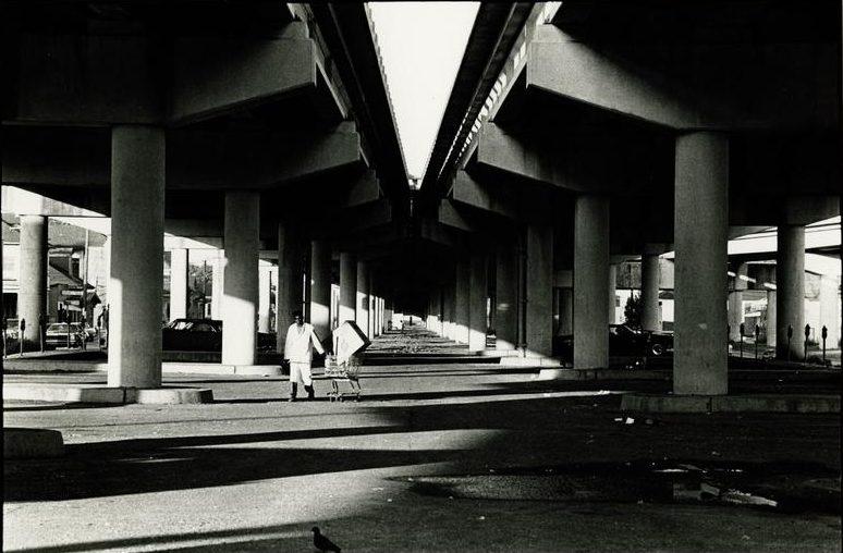Black and white photo of two people walking towards the camera under a large, symmetrical overpass. Sunlight creates stark shadows and patterns on the ground. In the background, cars and buildings are partially visible.