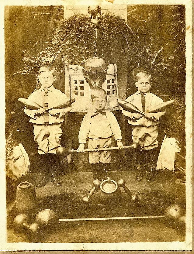 Antique sepia photograph of three young boys standing with exercise equipment, including dumbbells and Indian clubs, in front of a backdrop featuring greenery and a house. The boys wear early 20th-century attire with ties and shorts.