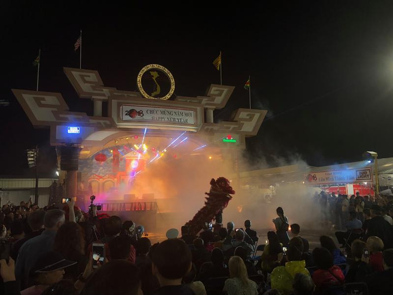 A vibrant night scene at a festival with a dragon dance performance on stage. Bright lights and smoke add to the spectacle as a crowd watches. The stage is decorated for the event, and the scene is bustling with excitement.
