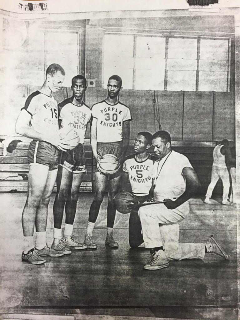 Black and white photo of a basketball team in a gym. Four players in Purple Knights jerseys and shorts stand holding a basketball. A coach kneels beside them with another basketball, wearing a T-shirt and pants. Gym equipment is visible in the background.